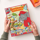 Titelblatt des Kindermagazins „Benjamin Blümchen“ mit der gezeichneten Elefantenfigur in roter Kleidung und roter Kappe. Im Hintergrund sind bunte Illustrationen eines grünen Chamäleons, einer Lupe, einer Rechenpyramide und einer Wüstenlandschaft zu sehen. Textelemente: „Gemüse-Snacks“, „So sehen Tiere“, „Ein fliegendes Chamäleon“, „Doras Rechenpyramide“ und „Wissen“. Zwei Hände halten das Heft auf einem hellen Tisch.