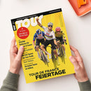 Titelblatt des Magazins „Tour“ mit gelbem Hintergrund, im Zentrum drei männliche Radsportler in Rennkleidung auf Fahrrädern, der mittlere hebt den rechten Arm. Linke obere Ecke mit rotem Kreis und Text „+ 64 Seiten! Extra-Heft zur Tour de France“. Weitere sichtbare Texte: „Tour de France Feiertage“, „Günstige Radhosen“, „Team Telekom“, „Die besten Touren am Arlberg“. Zwei Hände halten das Heft über einem hellen Tisch.