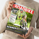 Titelbild des Magazins „ROADBIKE“ mit einem männlichen Rennradfahrer in hellgrünem Trikot, weißem Helm und Sonnenbrille auf weißem Rennrad, aufgenommen in Bewegung auf sonniger Straße vor grünem Hintergrund. Textaufschriften u. a.: „ENDURANCE RENNER“, „Kettenöle – 13 Schmierstoffe im Test“, „Gravel-Racer“, „ProfiSport“, „Fitness“, Hinweise zu Rädern, Ausstattung und Kaufberatung. Hände in beigem Pullover halten das Magazin.