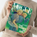Person in beigem Strickpullover hält ein Magazin mit dem Titel „VOGUE Deutsch Collector’s Issue“. Auf dem Cover sind mehrere kaleidoskopartig gespiegelte Ansichten einer weiblichen Person mit blondem Haar in einem blau-grünen gemusterten Kleid zu sehen. Sichtbare Texte: „MALA EMDE“, „THE WOMEN WE ARE“, „EIN KALEIDOSKOP DES FRAUSEINS“. Hintergrund in kräftigem Grün mit geometrischen Spiegelungen.