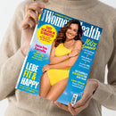Person in beigefarbenem Strickpullover hält eine Ausgabe des Magazins „Women’s Health“ mit blauem Hintergrund und einer Frau in gelbem Badeanzug auf dem Cover; sichtbare Texte: „Easy schlank in 15 Minuten“, „Jana Ina Zarrella – Mit 50 beginnt für mich die beste Zeit“, „Lebe fit & happy“, „100 % Sommer“, „Glow-Guide für schöne Haut“, „Love-Check“, „Die schönste Beachwear für jede Figur“.
