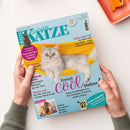Titelbild der Zeitschrift „Geliebte Katze“, Ausgabe Juli 2026, mit hellgrauer Katze in gelber Hängematte vor himmelblauem Hintergrund. Textaufschriften u. a.: „Einfach cool bleiben!“, „Tapfere Katze Lini – Sie sollte nicht mehr leben – heute erklimmt sie Berge“, „News und Tipps von Katzen-Experten“ und „Großer Ratgeber Balkonblumen“. Eine Person mit grünem Pullover hält das Magazin, im Hintergrund orangefarbene Schale.