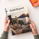 Magazin „Robb Report“ mit dem Titelthema „Luxus der Regeneration“ in Händen haltend; auf dem Cover eine hell gekleidete, männliche Person steht auf einer Terrasse vor einem See mit Bergen bei Sonnenuntergang, im Vordergrund sitzt eine weitere Person auf einem Sofa neben einem Glastisch mit Getränken; sichtbare Textzeilen u. a. „BODY & MIND ISSUE“, „Design. Stil. Reise. Motoren. Werte.“ und Ausgabeangabe „Juli/Aug 26“ am linken Rand.