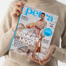 Person in beigem Wollpullover hält eine Ausgabe des Magazins „petra“ mit dem Titel „Die neue Sommermode“. Auf dem Cover steht eine Frau im gestreiften, hellen Kleid mit weitem Gürtel und Sandalen auf hellem Sandboden. Weitere sichtbare Texte: „Stylishe Beach-Bags“, „Lässige Kleider“, „Schöne Bade-Outfits“, „Coole Sonnenbrillen“, „Die besten Supplements“ und „Endlich Zeit zum Lesen“.