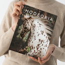 Magazin mit dem Titel „HOOFORIA“ in weißen Großbuchstaben, darunter kleinere grüne Schrift „Nature · Lifestyle · Western“. Auf dem Cover ein geschecktes Pferd mit braun-weißer Fellzeichnung und wehender Mähne vor verschwommenem Waldhintergrund. Unten links grünes Logo mit den Buchstaben „HC“ und dem Zusatz „VOLUME 2“. Gehalten von einer Person in beigem Strickpullover vor neutralem Hintergrund.