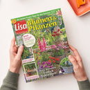 Zwei Hände halten eine Ausgabe des Magazins „Lisa Blumen & Pflanzen“ (August 2026) mit grün-gelbem Titeltext. Das Cover zeigt einen sommerlichen Garten mit pinken, roten und violetten Blüten in Kübeln und Beeten. Sichtbare Schlagzeilen: „Jetzt freuen wir uns auf… fantasievolle Miniatur-Welten“, „Endlich Dahlienzeit!“, „Erfrischende Wasserspiele“. Hintergrund unscharf, Oberfläche matt, im oberen rechten Bildbereich orangefarbene Schale.