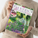 Frau in beigefarbenem Strickpullover hält ein Magazin mit dem Titel „Garten & Wohnen Träume“ (Ausgabe 3/2026). Auf dem Cover ein farbenfroher Garten mit Blumenbeeten in Lila-, Rosa- und Grüntönen, einer weißen Gartenbank im Hintergrund und dem Schriftzug „Willkommen im Sommergarten“. Oben links rundes Bild einer Kirschtorte mit der Aufschrift „Köstliches mit Kirschen“.