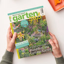 Titelseite der Gartenzeitschrift „gartenspaß“ vom Juli 2026 mit grün-gelbem Logo und Titelzeile „Blütenfest im Cottage-Garten“. Hauptfoto zeigt einen bunten, dicht bepflanzten Garten mit Holztisch, vier bunten Stühlen und Blüten in Rosa, Lila und Gelb. Seitlich kleinere Bilder und Texte zu Themen wie „Süße Sommerfrüchte“, „Tolle Taglilien“ und „Schritt für Schritt – Nisthilfe für Wildbienen bauen“. Zwei Hände halten das Magazin über hellem Tisch.