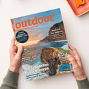 Titelbild der Zeitschrift „outdoor“ mit orangefarbenem Logo und Unterzeile „WANDERN. REISEN. ABENTEUER.“; zeigt eine steile Küstenlandschaft mit einer Person in gelber Jacke auf einer Klippe über dem Meer bei Sonnenuntergang. Text auf dem Cover: „Die schönsten Küstentouren“, „Peaks of the Balkans“, „Zelte im Sturm“ und „Die besten Produkte 2026“. Zwei Hände halten das Magazin über einer hellen Tischfläche mit orangefarbenem Gegenstand.