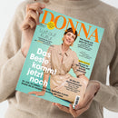Titelseite der Zeitschrift „DONNA“ mit türkisfarbenem Hintergrund, orangem Logo-Schriftzug oben und einer lächelnden Frau in beigefarbener Bluse und glitzerndem Rock in Sitzposition. Textaufschriften u. a.: „Das Beste kommt jetzt noch“, „Lust auf Kultur“, „Hier bade ich ganz tief mich in Deutschland“, „Neue Frisur“, „Ist das ein Hormonbauch?“. Gehalten von einer Person in beigefarbenem Strickpullover vor hellem Hintergrund.