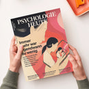 Magazin „Psychologie Heute“, Ausgabe August 2023, wird von zwei Händen mit grünem Strickärmel gehalten. Titelgrafik zeigt eine gezeichnete Person mit dunklem Haar in Profilansicht, die ein Tablet hält, das ihr eigenes Spiegelbild mehrfach wiederholt; Hintergrund in Rosa‑, Rot‑ und Beigetönen. Sichtbare Texte: „Immer mehr war irgendwann zu wenig“, „Serien, Social Media, shoppen…“, „Glück beim Warten“, „Staunen und Ehrfurcht“, „Ozeanisches Gefühl“.