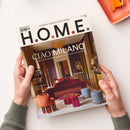 Titelblatt der Designzeitschrift „H.O.M.E.“ mit der Überschrift „CIAO MILANO“ und dem Untertitel „Das Beste vom Salone del Mobile und der Milan Design Week“, abgebildet sind ein Wohnraum mit violett-goldenen Wänden, rotem Sofa, runden Hockern in Grau und Orange, ein goldener Kronleuchter und dekorative Spiegel; gehalten von zwei Händen über heller Tischfläche mit orangefarbener Schale oben rechts.