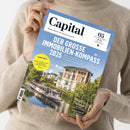 Person in beigefarbenem Strickpullover hält ein Exemplar der Zeitschrift „Capital“ mit Titel „DER GROSSE IMMOBILIEN-KOMPASS 2025“ vor hellem Hintergrund; Umschlag zeigt eine Flusslandschaft mit weiß-rotem Ausflugsboot, grünen Bäumen und mehrstöckiger Stadtvilla; sichtbare Textelemente: gelber Kreis mit „TRUMP-CRASH“, Untertitel zu Mieten, Kaufpreisen und Metropolen sowie weitere Themen zu Aktien, Gold und Start-ups.