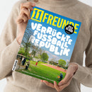 Person in beigem Strickpullover hält ein Magazin mit dem Titel „11 FREUNDE“ in gelber Schrift und der weißen Überschrift „VERRÜCKTE FUSSBALL REPUBLIK“ vor hellblauem Himmel; auf dem Cover ist ein Fußballplatz mit grüner Wiese, Laufbahn, Bäumen und mehreren spielenden Personen zu sehen, unten der Text „Eine Reise zu den kuriosesten Plätzen des Landes“, rechts oben ein schwarzer Kreis mit der Aufschrift „SPIELER DER SAISON 2024 / 25 Wahl“.