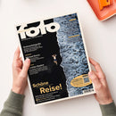Zwei Hände halten die Augustausgabe 2026 des „fotoMAGAZIN“ mit dem Untertitel „Die ganze Welt der Fotografie“. Titelbild zeigt eine Luftaufnahme einer Person in dunkler Badebekleidung am Rand von Meeresschaum und dunklem Wasser. Aufschriften: „Drohnenfotografie“, „Michael Kenna“, „Leichtes Gepäck“, „Schöne Reise!“, „Im Test: Systemkameras“ und „Smartphone Oppo Find X3 Ultra“.