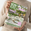 Person in beigefarbenem Strickpullover hält eine Ausgabe der Zeitschrift „WOHNIDEE“ mit grünem Titelkopf und Unterzeile „Wir lieben unser Zuhause!“. Auf dem Cover ein sommerlich gedeckter Gartentisch mit grün-weißer Dekoration, Glasgeschirr und Blüten, flankiert von Texten wie „Grüne Wohlfühldeko“, „Hitzeschutz“ und „Kleine Fläche, viel Sommer!“. Im Hintergrund blühender Flieder und grüne Pflanzen unter Lichterkette.