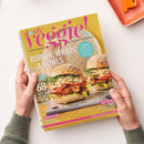 Gleichmäßig beleuchtetes Titelbild der Zeitschrift „slowly veggie! Vegetarisch & vegan genießen“ in gelb-pinkem Design, gehalten von zwei Händen vor hellem Hintergrund; mittig zwei Sesam-Burger mit Gemüse und Soße auf grauer Platte, Text u. a.: „Burger, Wraps & Bowls“, „68 Gute-Laune-Rezepte für den Sommer“, „Smokey Jackfruit-Burger mit Coleslaw“, „Coolcation“ und „Beerensaison“.