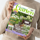 Person in beigem Strickpullover hält mit beiden Händen das Magazin „mein schöner Garten“, Ausgabe Juli 2026. Das Titelbild zeigt eine sommerliche Gartenszene mit weißen Blüten, Sitzbank, Holztisch und grüner Bepflanzung. Aufschriften u. a.: „Weiße Blüten im Sommergarten“, „Ideen mit Wasser“, „Jetzt schon ernten!“, „Alles Wichtige in diesem Monat“, Website „mein-schoener-garten.de“.