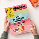 Titelblatt des Magazins „ZEIT WISSEN“ mit gelbem Feld links und der Aufschrift „DER MOND – Die große Kraft am Himmel und ihr Einfluss auf die Menschen“. Haupttitel in schwarzen Großbuchstaben: „JETZT IST ERST MAL SOMMER – Wie man sich von Sorgen frei macht. Und neue Lebenslust gewinnt“. Illustration zeigt zwei Personen auf Liegestühlen an einem rosa Strand mit Blick aufs Meer. Hände halten die Zeitschrift vor hellem Hintergrund.