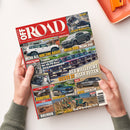 Titelbild des Magazins „OFF ROAD – Das 4x4-Magazin für die Freiheit auf Rädern“ mit mehreren Fahrzeugfotos, darunter Pick-ups und Geländewagen. Textelemente wie „Toyota Land Cruiser 6-Zyl.-Motor“, „MEHR ALS NUR EINE BASIS“, „DER VIELLEICHT BESTE J ALLER ZEITEN“, „TEST HONDA CR-V“ und „BOLIVIEN“ sind sichtbar. Das Heft wird von zwei Händen mit grünem Strickpullover auf hellem Tisch gehalten, daneben ein orangefarbenes Tablett.