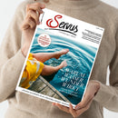 Person in beigefarbenem Strickpullover hält eine Ausgabe der Zeitschrift „Servus – einfach gut leben“ in beiden Händen; das Titelbild zeigt Beine einer Person mit gelbem Rock, die ihre Füße in klares Wasser eines Sees taucht, neben einem hölzernen Steg. Sichtbare Texte: „Warum Wasser Wunder wirkt – Gesunde Rituale und quellfrische Erkenntnisse“ und roter Kreis mit „So schmeckt die Kindheit – Eine Liebeserklärung an den Marillenstrudel“.