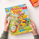 Kinderzeitschrift mit dem Titel „Disney Winnie Puuh“, hellblauer Hintergrund und bunte Zeichentrickfiguren: Winnie Puuh, Tigger, Rabbit, Ferkel und I-Aah auf einer Wiese mit Bäumen. Textfelder: „EXTRA: Shopping-Set 8 Teile“, „Poster und Rätsel“, „Naturforscher süßer Tiernachwuchs“, „Entdecken Malen Spielen“ und „Zum Herausnehmen!“. Zwei Hände halten das Heft über hellem Tisch mit orangefarbenem Tablett.