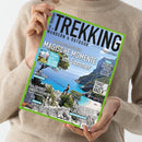 Person in beigefarbenem Pullover hält das Magazin „Trekking – Wandern & Outdoor“ mit grünem Rand und blauem Titelhintergrund. Haupttitel: „Magische Momente im Sommer“. Sichtbare Schlagzeilen zu Themen wie Allgäu, Wolf Trail, Salzburger Spaziergang, Sardiniens Küstenpfade, Klettern und Approachschuhe. Coverfoto zeigt eine Person auf Felsen über türkisblauer Bucht mit Klippen und sonnigem Himmel.