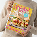 Person in beigefarbenem Strickpullover hält das Magazin „slowly veggie!“ mit gelbem Hintergrund, pinkfarbenem Logo und der Unterzeile „vegetarisch & vegan genießen“ in der Hand. Auf dem Cover befinden sich zwei Burger mit Sesambrötchen, Gemüse und Coleslaw, dazu Texte wie „Burger, Wraps & Bowls“, „68 Gute-Laune-Rezepte für den Sommer“, „Beerensaison“ und „Coolcation“.