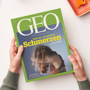 Titelblatt der Zeitschrift „GEO“ mit grün-blauem Hintergrund, großem weißem „GEO“-Logo und gelbem Haupttitel „Hilfe bei chronischen Schmerzen – Neue Therapien für Körper und Seele“. Darunter eine verschwommen dargestellte weibliche Person mit gesenktem Kopf und Händen am Kopf. Weitere Texte: „Psychosomatik“, „Ghana“ und „Indonesien“. Gehalten von zwei Händen über hellem Tisch mit orangefarbener Schale am Rand.