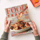 Titelbild der Zeitschrift „Food and Travel“ in deutscher Ausgabe, gehalten von zwei Händen vor hellem Hintergrund. Abgebildet ist eine Schüssel mit gebratenen Garnelen, eine Garnele wird mit Essstäbchen gehalten. Sichtbare Texte: „Asiatische Küche“, „Rezepte von der Donau“, „City Guide München“, „Spektakuläre Spas“, „Afrikas Küsten“ und „Foodie-Hotspots – Städte zwischen Streetfood und Sternen“.