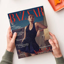 Magazin „Harper’s Bazaar“ mit rotem Logo auf dunkelblauem Hintergrund, gehalten von zwei Händen über einer hellen Fläche. Auf dem Cover steht eine weibliche Person in schwarzem, strukturiertem Kleid vor einem sandfarbenen Hintergrund. Textelemente: „MODE – Sonnengelb, Supershort und Schmuck zum Verlieben“, „123 Gründe für Freude“, „Peloponnes“, „Star-Architekt Bjarke Ingels“, „GNTM-Gewinnerin AuRélie“.
