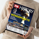 Helles Magazincover „P.M. HISTORY“ mit dem Titel „WENDEPUNKTE – 80 Jahre, die die Welt veränderten“, Unterzeile „Von Hiroshima bis zum iPhone“. Abgebildet sind ein Atompilz, ein Astronaut, das Brandenburger Tor mit Menschenmenge und eine iPhone-Grafik. Weitere Textfelder unten: „SAMT & SEIDE“, „HALLO?!“ und „WM-FIEBER“. Gehalten von einer stehenden Person in beigem Strickpullover vor neutralem Hintergrund.