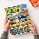 Titelblatt des Reisemobil-Magazins „promobil“ mit gelbem Schriftzug auf blauem Hintergrund. Im Mittelpunkt ein weißes Wohnmobil auf einer Wiese, davor zwei Menschen auf Campingstühlen. Überschriften: „Schlank und günstig“, „14 Campingbusse für große Menschen“, „Solartaschen im Test“, „Relaxen mit Seeblick“ und „Westfalia James Cook“. Gehalten von zwei Händen über einem hellen Tisch mit oranger Ablageschale.