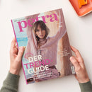 Titelbild der Lifestyle-Zeitschrift „petra“, gehalten von zwei Händen über einer hellen Tischfläche neben einer orangefarbenen Schale. Auf dem Cover eine Frau in einem hellrosa Zopfpullover und beiger Hose vor sandfarbener Steinwand. Sichtbare Texte: „DER TREND-GUIDE“, „Traumkleider +++ IT-Bags +++ Summer-Shoes +++ Beauty-Highlights“, „BEST OF GERMANY“, „Stil & Haltung“, „Mein Leben! Mein Luxus!“.
