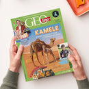 Titelblatt der Kinderzeitschrift „GEOlino“ mit grünem Kopfbereich und großem Schriftzug „GEOlino“. Im Zentrum ein Foto zweier Kamele in einer Wüstenlandschaft unter der Überschrift „Kamele – Was die genügsamen Tiere zu Wüstenprofis macht“. Links oben ein kleiner männlicher Moderator neben der Aufschrift „Checker Tobi – Filmtiere“, rechts unten ein Junge mit Mikrofon und Textfeld „Volltreffer! Der Nachwuchsreporter …“. Logo von UNICEF unten rechts.
