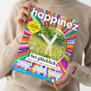 Eine Person in beigefarbenem Strickpullover hält die Zeitschrift „happinez – Das Mindstyle Magazin“. Das Cover zeigt eine bunte, kreisförmige Illustration eines Menschen beim Handstand auf einer Wiese mit Blumen, umgeben von farbenfrohen Strahlen. Sichtbare Texte: „Sei glücklich – Alles strahlt heller, wenn Du lachst“, „Selbst-Erkenntnis“, „Liebes-Magie“, „Aktive Hoffnung“, „Weiblichkeit – Die Superkraft Deines Zyklus“, gelbes Feld: „EXTRA Zauberhafte Lesezeichen“.