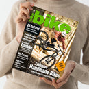 Person in beigefarbenem Strickpullover hält die August-2023-Ausgabe des Magazins „bike“. Auf dem Cover: eine männlich gelesene Person mit weißem Helm, rotem Hemd und schwarzem Fahrrad in dynamischer Fahrhaltung im Wald. Auffällige grüne Titelaufschrift „bike“, weitere sichtbare Texte: „26 Zoll lebt“, „Exklusive Handmade-Bikes“, „Rund um die Eurobike“, „Power-Motoren“, „Mr. Canyon“ und „High-Flow-Ventile“.