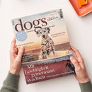 Titelblatt des Magazins „dogs“ mit großem Schriftzug und Foto eines Dalmatiners vor Sonnenuntergang am Meer. Sichtbare Texte: „Mit Leichtigkeit gemeinsam wachsen – Mehr Freude in der Erziehung“, „Reif für die Insel – Sardinien mit Hund“, „Hilfe für die Seele“, „GUT GEMACHT!“, sowie blauer Kreis mit „EXTRA – Die Welt aus der Sicht des Hundes“. Gehalten von zwei Händen vor hellem Untergrund, orangefarbenes Tablett im Hintergrund.