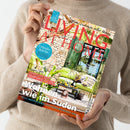 Eine hellhäutige Person in beigem Wollpullover hält eine Ausgabe der Zeitschrift „LIVING AT HOME“ mit dem Titel „Wohnen wie im Süden“. Auf dem Cover: eine mediterrane Steinfassade mit grün lackierter Holztür, davor grüne Gartensofas, Pflanzen und Wohnaccessoires. Sichtbare Schriftzüge: „Urlaubsküche“, „Sommerliche Ideen für den Tisch“, „Citytrip München“ und „Tolle Inspirationen von Palma bis Buenos Aires“.