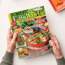 Magazin „meine gute Landküche“ mit grün-gelbem Titelkopf und der Aufschrift „Genießen im Sommer“ auf dem Cover, gehalten von zwei Händen über hellem Hintergrund. Abgebildet sind ein roter Auflaufform mit Gemüse-Quiche, daneben weitere Speisen wie Gebäck, Steaks und Kuchenstücke. Zusätzliche Texte: „Hausgemacht schmeckt’s am besten!“, „Ländlicher Genuss & regionale Vielfalt“, „Kräuter pflegen & Tipps zum Würzen“.