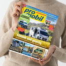 Eine Person in beigefarbenem Pullover hält die Zeitschrift „promobil“ in den Händen. Das Titelbild zeigt einen weißen Campingbus auf einer Wiese mit sitzender Person daneben, darüber die gelbe Überschrift „Schlank und günstig“. Weitere Textfelder: „Kauf-Tipp: 14 Campingbusse für große Menschen“, „Solar­taschen im Test“, „Relaxen mit Seeblick“, „Westfalia James Cook“. Die Farbgestaltung ist gelb, blau und rot auf hellem Hintergrund.