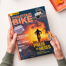 Titelblatt der Zeitschrift „mountainBIKE“ mit großem orangefarbenem Logo oben links, Untertitel „ALLES IST FAHRBAR“ und Fotomotiv eines Mountainbikers vor orange-gelbem Sonnenuntergang. Mehrere Textfelder: „Der PREIS ist HEISS“, „21 Enduro- & Trail-Reifen im Test“, „Orbea – Das neue E-MTB-Flaggschiff“, runder Aufkleber „Neu – 16 Seiten Community“. Oben rechts Fahrradschaltung mit Schriftzug „Shimano XT Di2 im Stresstest“. Hände halten das Magazin über hellem Tisch.