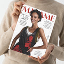 Magazincover mit rotem Schriftzug „MADAME“ im oberen Bereich, zentrale Abbildung einer lächelnden weiblich erscheinenden Person mit kurzem, dunklem Haar, weißem Shirt und schwarzem Kleid mit roten Blumenstickereien. Sichtbare Texte: „PURE JOY!“, „Die schönsten Aussichten auf die kommende Modesaison“, „REISE ZU DEN STERNEN“, „BEST OF DESIGN Neue Möbel aus Mailand & Paris“. Hände halten das Magazin vor beigem Pulloverhintergrund.