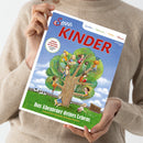 Person in beigem Strickpullover hält ein Magazin mit dem Titel „Servus Kinder“. Das Cover zeigt eine farbenfrohe Illustration eines großen Baumes mit mehreren spielenden und lesenden Kindern, einer Wiese mit Kuh und Zaun im Hintergrund. Oben stehen die Wörter „Spielen“, „Staunen“, „Raten“, „Wissen“, unten der Schriftzug „Das Abenteuer deines Lebens – Tausend Ideen für große und kleine Entdecker“.