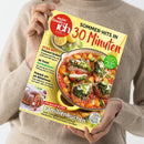 Person in beigefarbenem Strickpullover hält ein Magazin mit dem Titel „meine Familie & ich“ in der Ausgabe 8/2026, Untertitel „SOMMER-HITS IN 30 Minuten“. Auf dem Cover ein gelber Topf mit Hähnchen, Gnocchi, Tomaten und Kräutern, daneben Zitronenscheiben. Textausschnitte: „EXTRA-HEFT Preiswert kochen“, „Im Trend: Gartenkräuter“, „Familienkuchen – Mit Beeren & Obst“.