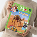 Person mit beigefarbenem Strickpullover hält das grün umrandete Kindermagazin „GEOlino“ mit Titelthema „KAMELE – Was die genügsamen Tiere zu Wüstenprofis macht“. Auf dem Cover stehen zwei Kamele in einer rötlichen Wüstenlandschaft, oben ein Porträt von „Checker Tobi“ mit rosa Schweinchen und dem Text „FILMTIERE“, unten ein gezeichneter Manga-Ausschnitt und rechts ein Kind mit Brille und Fußball.