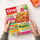 Magazin „tina – Koch & Back-Ideen“ Ausgabe Juli 2026, gehalten von zwei Händen über hellem Hintergrund; Titelbild mit großem Foto eines italienischen Auflaufs aus Tomaten, Hähnchen und Basilikumblättern, umgeben von Texten wie „Appetit auf Italien“, „Mehr als 100 Rezepte – mit Geling-Garantie“, „Süße Erfrischung: Himbeerschnitten“ und „Fix aufgetischt!“. Farbig gestaltetes Layout in Rot, Gelb, Türkis und Weiß.