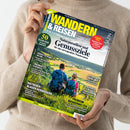 Person in beigem Strickpullover hält das Magazin „Wandern & Reisen – Ausgabe 03-26 Sommer“. Auf dem Titelbild sitzen zwei Personen mit blauen Rucksäcken in grüner Wiesenlandschaft und blicken auf ein weites Tal mit Feldern und Hügeln. Sichtbare Texte: „Naturparadiese und Genussziele“, „50 frische Ideen“, „Ratgeber: Blasen verhindern“, „Premium-Wanderwelten“ sowie weitere regionale Themen zu Wanderzielen in Deutschland und den Alpen.