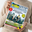 Person in beigefarbenem Strickpullover hält ein Magazin mit dem Titel „e‑bikeTOUREN MAGAZIN“ in den Händen; auf dem Cover zwei Personen mit Helmen auf E‑Bikes auf einem Waldweg. Sichtbare Texte: „20 Traumtouren für Genießer“, „Klassiker Donau‑Radweg“, „Märchenhafte Mittelgebirge“, „E‑Bike‑Motoren“, „Frühling/Sommer“, gelber Kreis mit „45 Ideen für den perfekten Urlaub“ und Hinweis „Ein Sonderheft von Bike & Travel“.