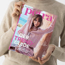 Person in beigem Strickpullover hält eine Ausgabe des Modemagazins „petra“ mit rosafarbenem Titelkopf. Auf dem Cover ist eine weibliche Person in einem rosafarbenen Zopfpullover mit passender Umhängetasche vor heller Felsenkulisse zu sehen. Lesbare Texte: „DER TREND-GUIDE“, „TRAUMKLEIDER +++ IT-BAGS +++ SUMMER-SHOES +++ BEAUTY-HIGHLIGHTS“, „BEST OF GERMANY“, „STIL & HALTUNG“ sowie „Mein Leben! Mein Luxus!“.